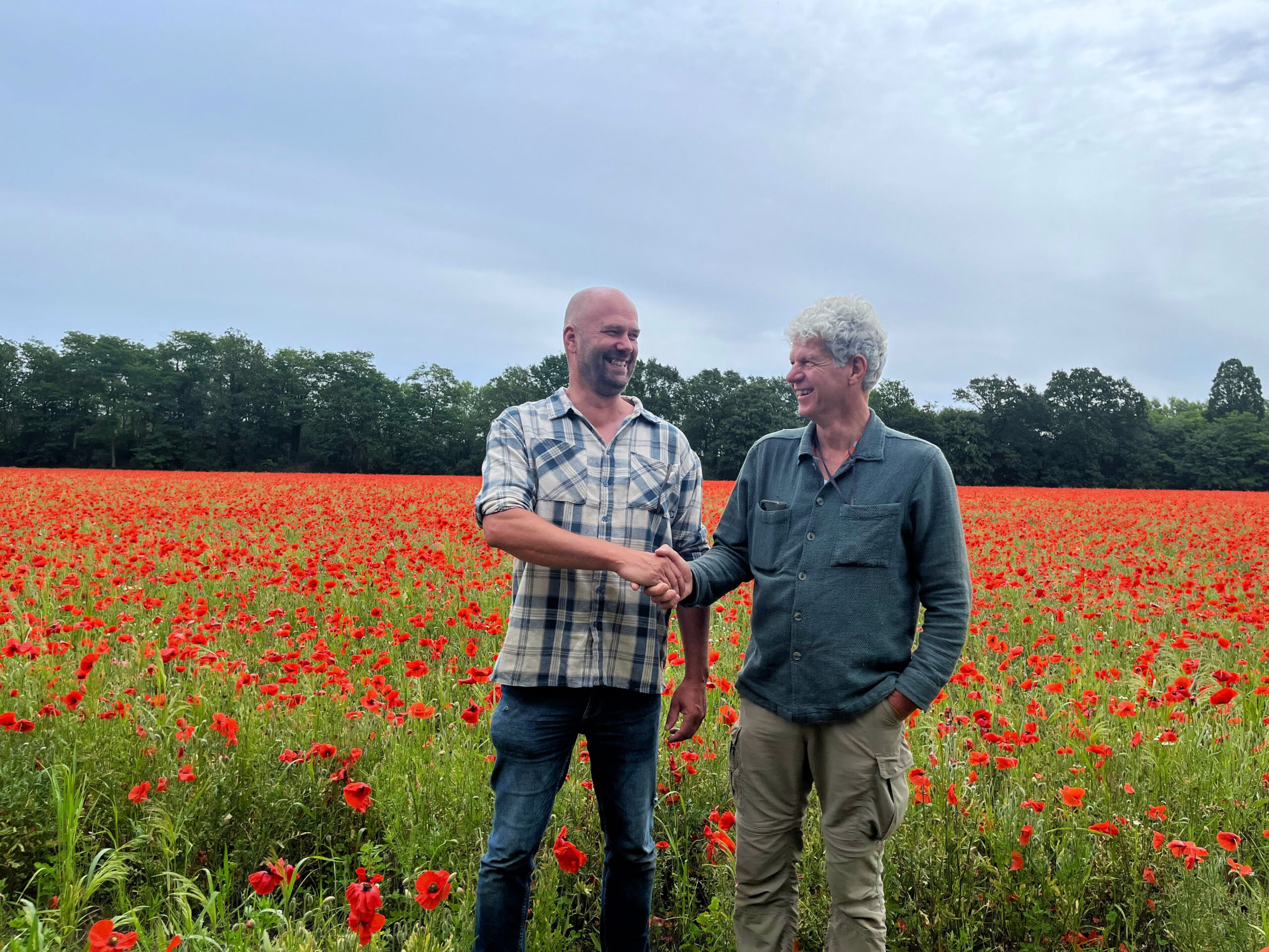 Egbert Peter Samen Egbert Hogendoorn en Peter de Groot schudden elkaar lachend de hand in een veld met bloeiende klaprozen.